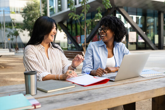 Female Students Studying Together Outdoors Using Laptop At University, having fun at MBA business uni campus