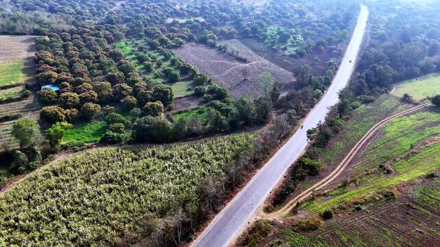 Drone shot capturing a sharp countryside road bend cutting between agricultural fields and woodland areas, with vehicles moving through layered rural landscapes and rugged terrain.