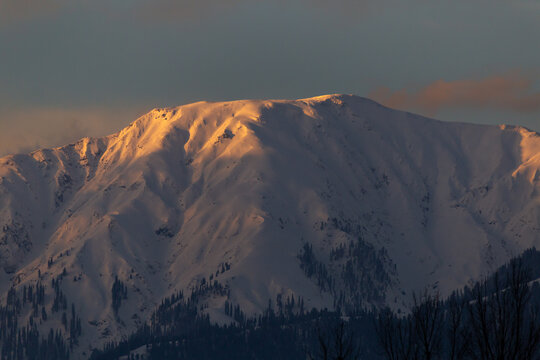 View of Makra Peak with snow-covered slopes and golden sunset light on the summit above dark evergreen trees in Balakot, Khyber Pakhtunkhwa, Pakistan.