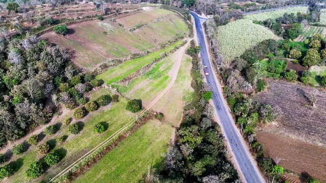 Wide cinematic aerial of a curving mountain road showcasing heavy vehicles traveling through forest-covered terrain, with layered greenery, dry hillsides, and dramatic winding roadway patterns from ab