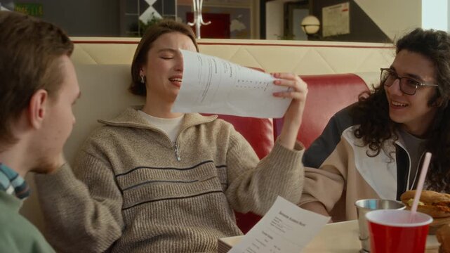 Medium shot of young Caucasian girl complaining to male friends about exam results while eating out together at fast food diner, crumpling score sheet, throwing away in frustration and disappointment