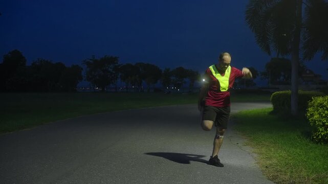 Middle-aged Asian man stretching quadriceps muscles after jogging at night with LED safety light