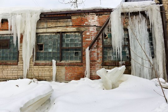 Giant icicles drape over a weathered brick industrial building facade with frosted glass block windows beneath a snowy roofline