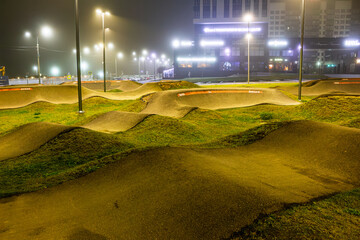 Naklejka premium Empty pump track in a city park at night, illuminated by bright glowing street lamps under a misty sky, creating a serene urban atmosphere for cycling sports: Russia - December 2, 2025