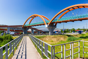 Fototapeta premium Modern arched pedestrian bridge crossing a busy highway, metal footbridge structure with vibrant orange arcs against a clear blue sky on a sunny summer day, urban infrastructure architecture
