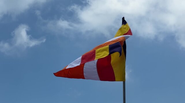 A colorful Buddhist flag moves freely in the wind on a tall pole against a bright blue sky with clouds forming a symbolic element of spiritual identity. Buddhist culture.