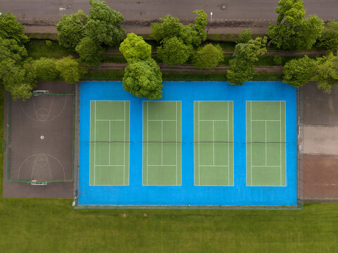 Aerial view of four green tennis courts on a vibrant blue surface next to a basketball court and lush green trees in Edinburgh, Scotland, United Kingdom.