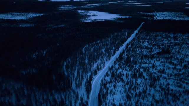 Winter road through boreal forest at blue hour, Lapland, Finland. Elevated drone view of a white road diagonal across dark snow laden conifers and frozen bog clearings under deep blue dark sky.