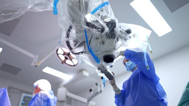 Overhead view of sterile draped surgical microscope being positioned by surgeon. Upward angle shows plastic drape with blue tape, surgical lamp at left, white ceiling.