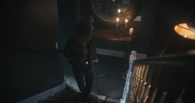 Young woman cautiously walking up stairs of scary, dark home then being distracted by something - high shot