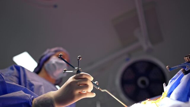Surgeon hand holding cranial fixation clamp during neurosurgery. Low angle, gloved hand grips metal clamp in foreground, blurred surgeon and surgical lamp in background.