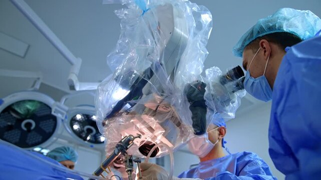 Neurosurgeon using draped microscope with surgical lamp above. Surgeon peers into eyepiece, assistant works below, circular lights visible at left.