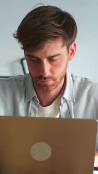 Concentrated young businessman with a beard using a laptop computer for remote work, pausing to think and look away thoughtfully before resuming his focused typing in a home office