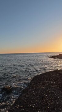 Gentle sea waves washing up on a rocky shore during a beautiful golden sunset, with the sun's reflection glittering on the calm water surface under a clear blue and orange sky