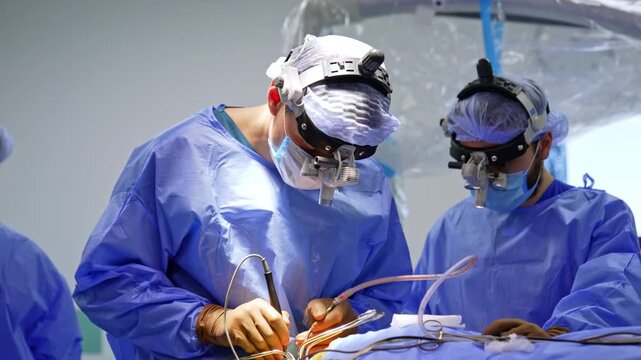 Two neurosurgeons with binocular loupes performing brain surgery. Both in blue gowns lean over operative field, instruments and suction tube held in gloved hands.
