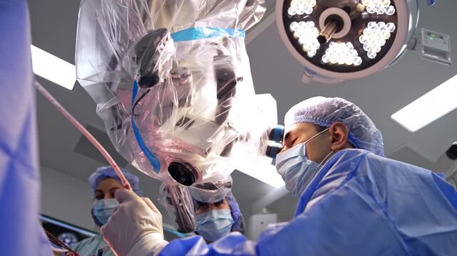 Neurosurgeon at draped microscope with LED surgical lamp. Surgeon peers into eyepiece, assistant holds tube, transparent plastic drape fills frame.