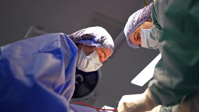 Two surgeons leaning over operative field during neurosurgery. Low angle upward view, blue and teal gowns, masks, caps, surgical lamp and drape visible above.