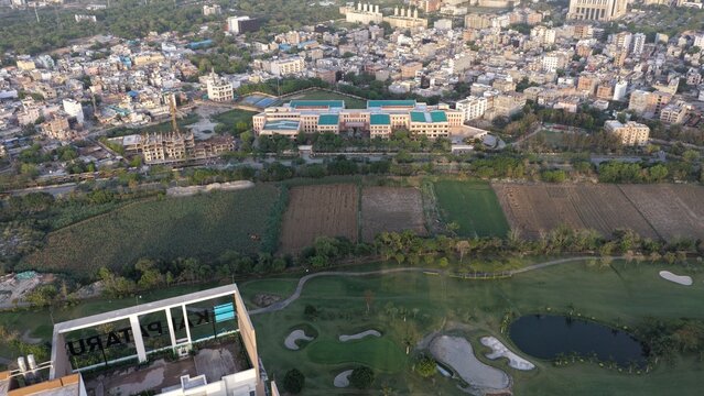 Aerial view of a lush green golf course, agricultural fields, and a large modern building complex with the urban skyline in the background Gurugram, Haryana, India.