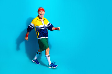 Obraz premium Active senior man in retro sportswear walking briskly against bright blue background, smiling while exercising and enjoying fitness and healthy lifestyle