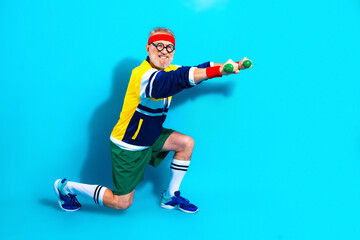 Obraz premium Senior man exercising with dumbbells in retro workout outfit on bright blue background, smiling and kneeling in a playful fitness pose promoting active lifestyle and joyful aging