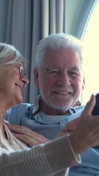 Affectionate elderly couple smiling and having fun while using a smartphone together, happily browsing the internet and enjoying their retirement sitting near a window at home