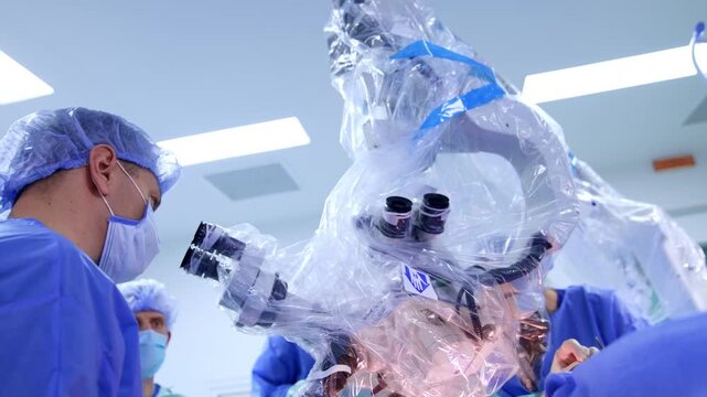 Surgeon looking into monocular microscope eyepiece during brain surgery. Low angle view, blue gown, draped microscope with blue tape, bright ceiling panels above.