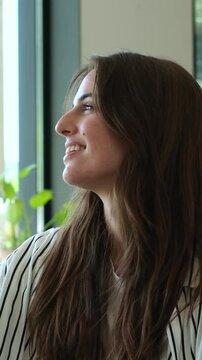 Young beautiful woman with long brown hair sitting by a window, smiling and enjoying the sunlight in a bright living room. Peaceful moment of a happy caucasian female relaxing at home