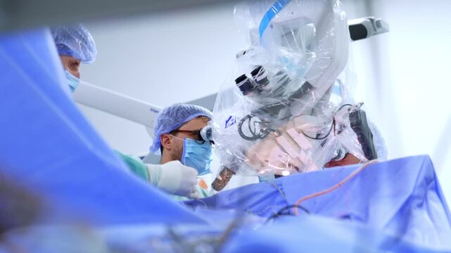 Neurosurgeon with safety glasses at draped microscope near blue drape. Surgeon leans to eyepiece, neuronavigation arm and assistant visible behind.