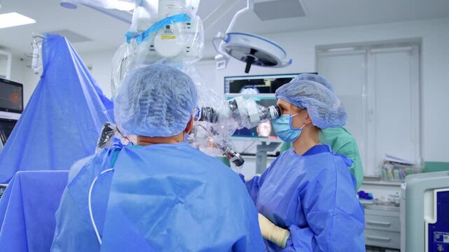 Two surgeons performing neurosurgery with microscope in operating room. Wide view of female and male in blue gowns using draped microscope, monitors and surgical lamps visible in background.