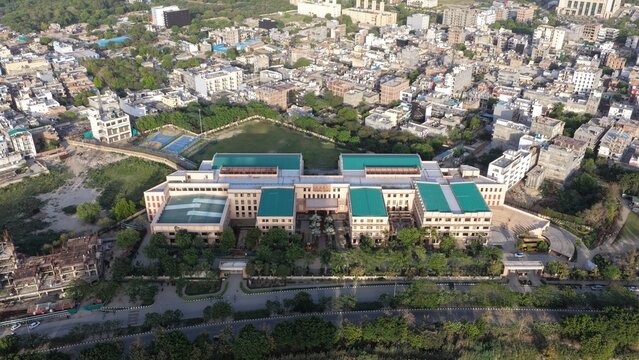 Aerial view of a large institutional building with green roofs and a sports field surrounded by dense urban housing and lush trees in Gurugram, Haryana, India.