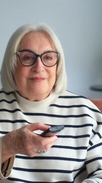 Cheerful elderly woman with gray hair and glasses sitting comfortably on a couch, holding a remote control and changing channels while watching a television show in her living room