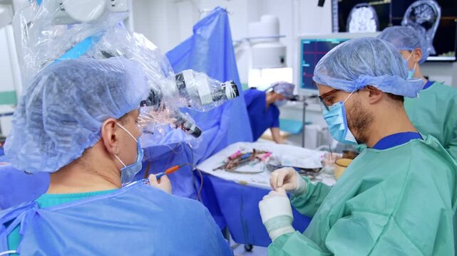 Neurosurgeon at microscope with assistant examining material during surgery. Surgeon in blue with loupe headband at eyepiece, teal gown assistant holds specimen at right.