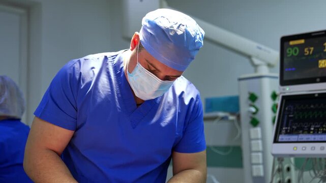 Neurosurgeon in scrubs leaning forward at operating table with vital monitors and anesthesia equipment visible. Male in blue cap and mask, patient monitor showing readings beside him.