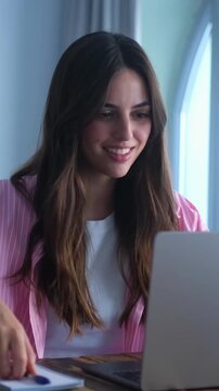 Focused young woman working on her laptop and taking notes in a notebook, smiling while studying online from home. The beautiful student enjoys remote education and online courses