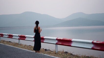 Fototapeta premium Young woman standing on road observing tranquil lake with distant mountains