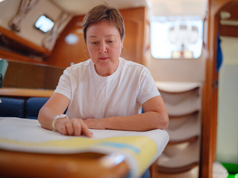 Mature woman sits inside yacht cabin analyzing paper sea chart for route planning. Navigation process involves calculating coordinates and path for upcoming sailing journey from Finike.