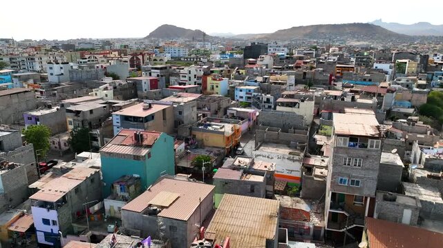 
Aerial footage of a vibrant hillside community in Cape Verde or a similar Atlantic coastal city. Brightly painted buildings cascade down a slope beside a major highway, with the ocean visible in the 