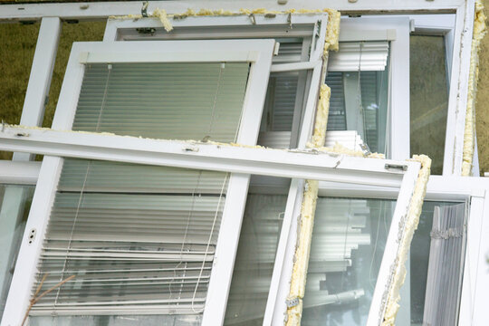 close up view of disassembled plastic windows stacked outside after being removed from a building with white frames glass window panes built in blinds and remaining yellow polyurethane foam