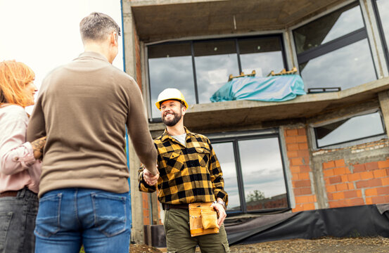Smiling construction worker shaking hands with an adult couple at a house construction site, celebrating a successful agreement.