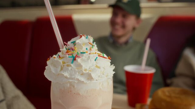 Close-up of delicious sweet milkshake with whipped cream, topped with sprinkles, in tall glass with straw served on table for group of zoomers chatting together at fast food restaurant