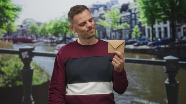 Man holding a brown envelope and showing a thumbs down gesture in a studio with canal backdrop and burgundy sweater; disappointment.