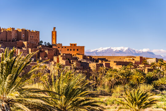 Visitors can see the Tifoultoute Kasbah fortress in Morocco along with palm trees and mountains in the background. The sun shines brightly on this historical site and nearby oasis.
