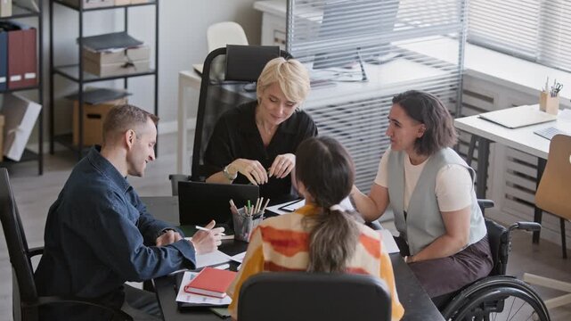Wide stab shot of managers and young female employee in wheelchair sitting at office table, discussing business plan and analyzing documents