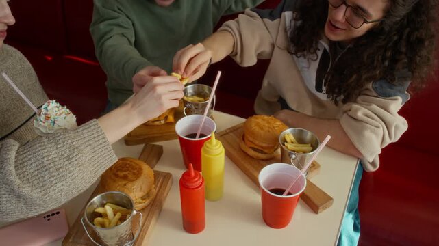 Medium high-angle shot of three joyous students toasting with fries, celebrating end of semester or school year at fast food restaurant, then tucking into tasty burgers with soda and milkshake
