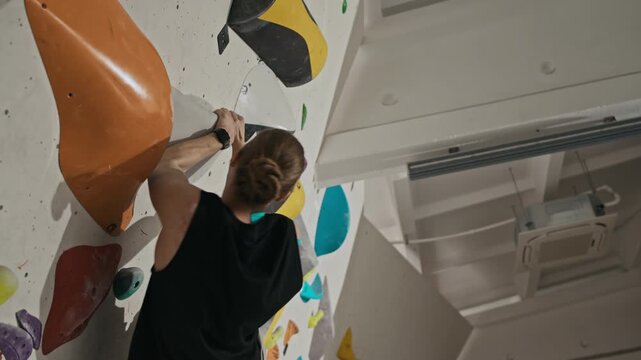 Young athletic man climbing indoor bouldering wall during workout, reaching for colorful holds and finding footing. Footage ideal for fitness brand ads or motivational sports content, low angle view
