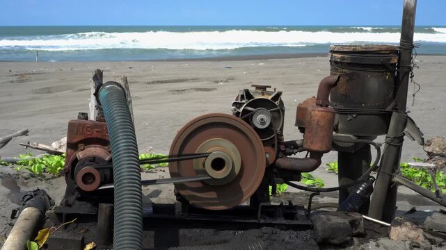 Static shot of an industrial water pump engine operating on a sandy beach with visible machine vibration, rotating wheel, and ocean waves in background.