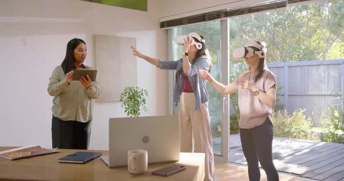 Asian trainer with tablet raising hand, speaking, training two women donning VR headsets in office