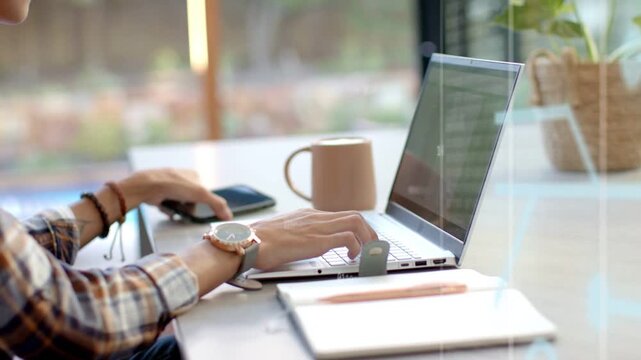 Man alternating typing on laptop, using phone at window desk, causing tech UI glow while working