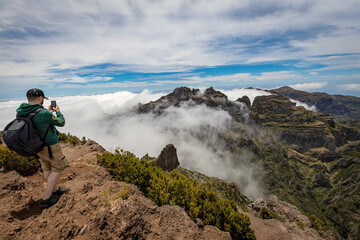 Fototapeta premium Man taking pictures on the Pico Ruivo summit, breathtaking view of the mountains covered with clouds