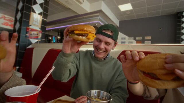 Medium dolly-in shot of three diverse cheerful zoomers, two guys and girl, toasting each other with hamburgers while eating out at retro style diner, celebrating successful exams at college
